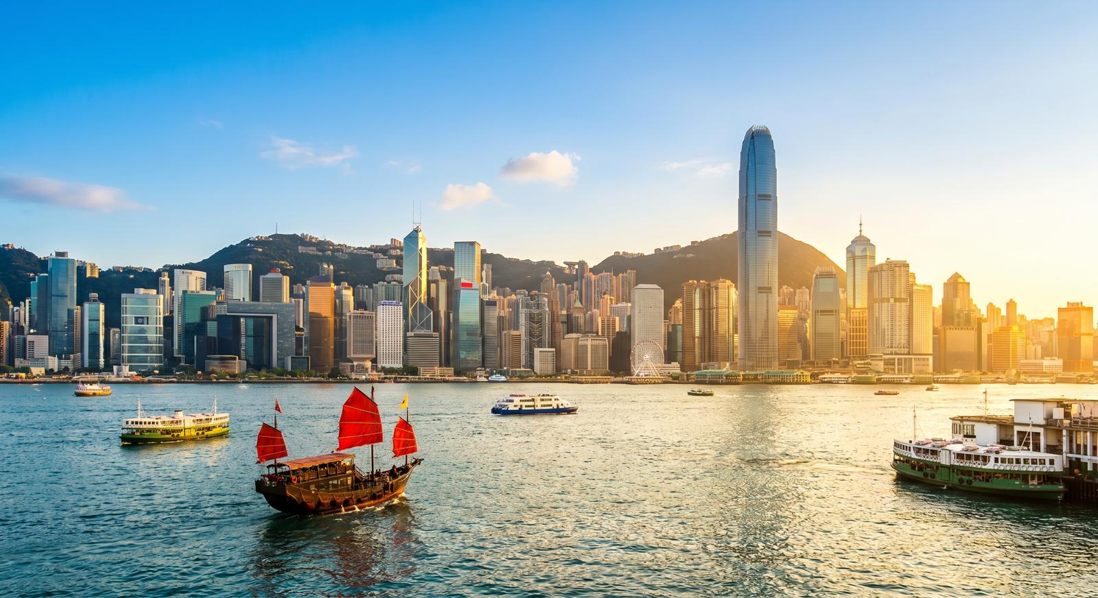 Panoramic city skyline and harbour in daylight with a red‑sailed traditional boat in the foreground